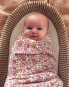 Baby swaddled in rose floral muslin lying in a woven bassinet