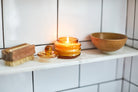 Lit Beam candle glowing amber on a tiled shelf beside a wooden brush and ceramic bowl