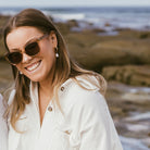 A woman smiling at a rocky beach, wearing the Lila Grace sunglasses