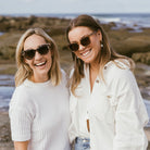 Two women at a rocky beach, both wearing Lila Grace sunglasses