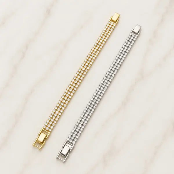 Two three-row cubic zirconia tennis bracelets, one gold tone and one silver tone, lying side by side on marble