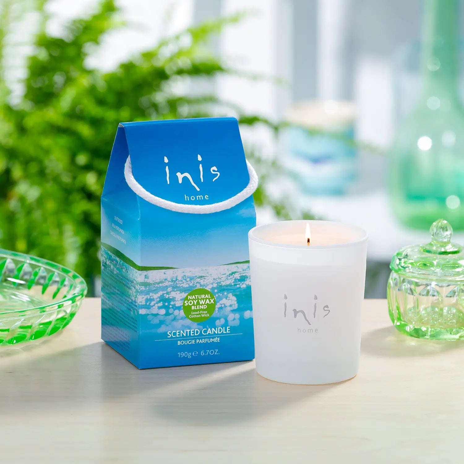 Lit Inis Scented Candle and its gift carton on a table with green glassware