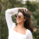 A laughing woman with one hand in her long dark hair, wearing the Cleo Bloom sunglasses outdoors