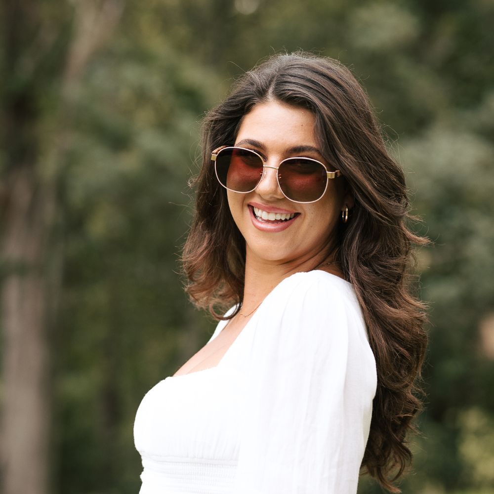 A smiling woman with long dark hair and a white top wearing the Cleo Bloom sunglasses outdoors, head turned to the side