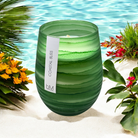 Wave Green Coastal Bliss candle on white sand beside turquoise water, framed by palm fronds and bright flowers