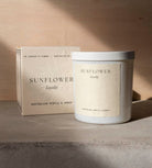 The Sunflower candle in its white vessel beside its cream box on a concrete ledge.