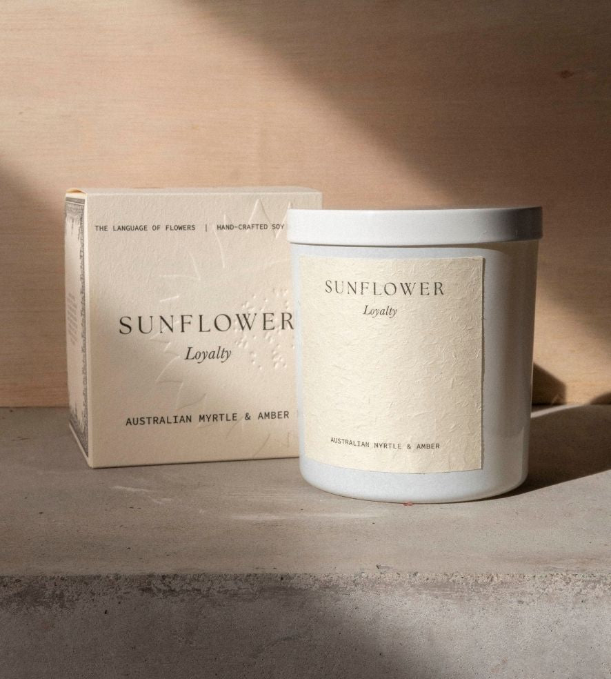 The Sunflower candle in its white vessel beside its cream box on a concrete ledge.
