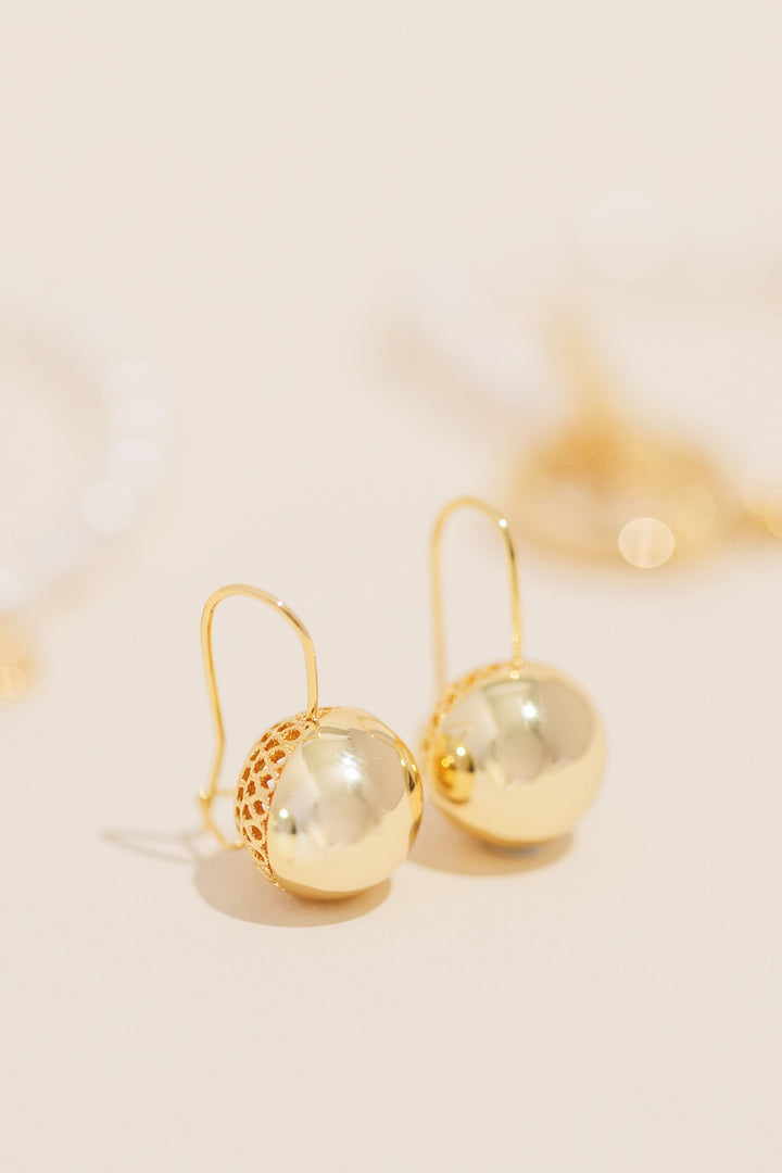 The Chelsea gold ball earrings photographed on a soft peach background.