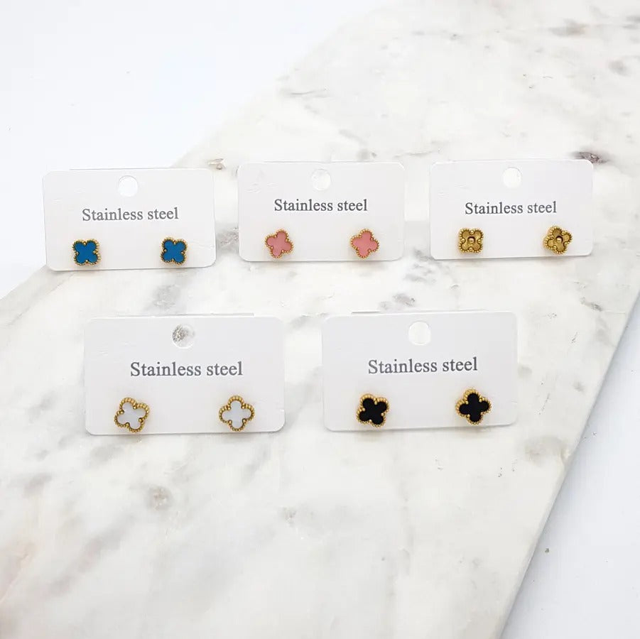Five pairs of small clover stud earrings on white cards — teal, pink, gold, white and black — laid on marble