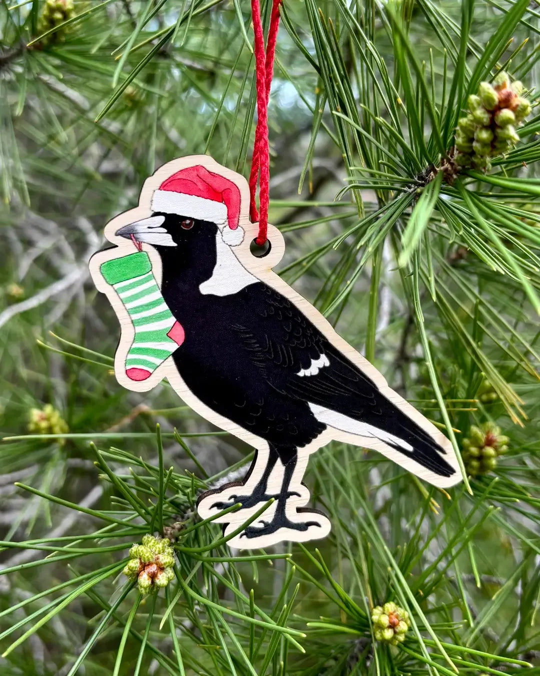 The wooden magpie decoration hanging outdoors from a pine branch, the magpie in a red Santa hat holding a striped stocking.