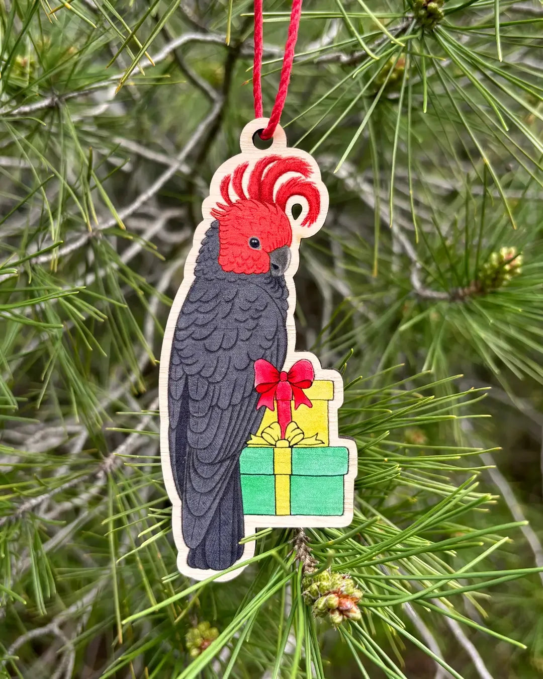 The wooden gang-gang cockatoo decoration hanging outdoors from a pine branch, showing its scarlet crest and the wrapped presents below.