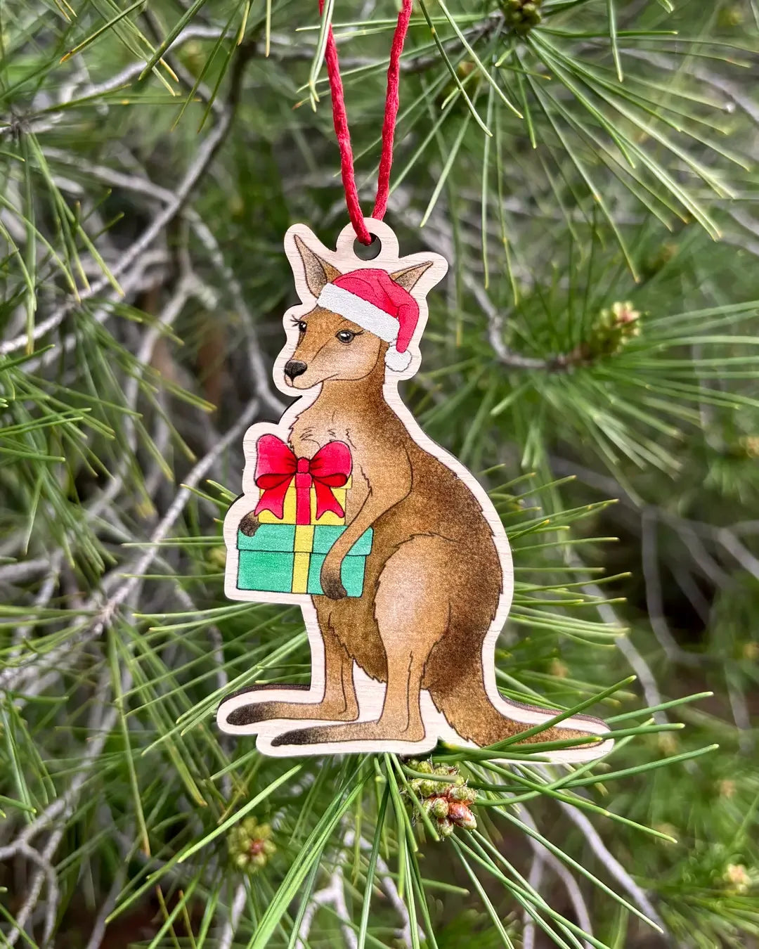 The wooden kangaroo decoration hanging outdoors from a pine branch, the kangaroo in a red Santa hat holding a wrapped present.