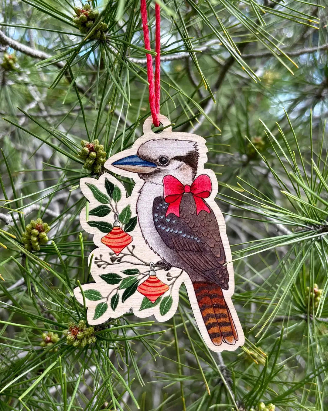 The wooden kookaburra decoration hanging outdoors from a pine branch, showing the red bow and the orange baubles on its leafy perch.
