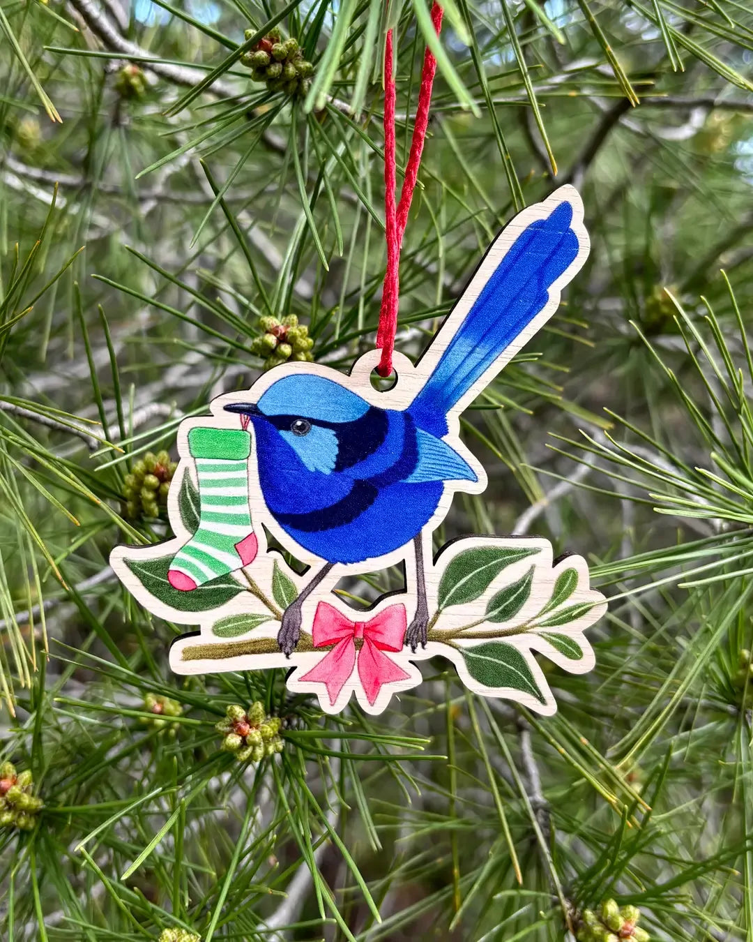 The wooden fairy wren decoration hanging outdoors from a pine branch, the blue wren beside a striped stocking and a pink blossom.
