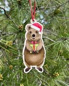 The wooden quokka decoration hanging outdoors from a pine branch, the quokka in a red Santa hat holding a small wrapped gift.