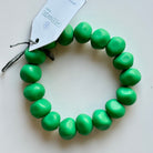 Rock bracelet in Emerald: a stretch bracelet of chunky emerald green resin beads with its swing tag and metal charm, laid on a pale surface.