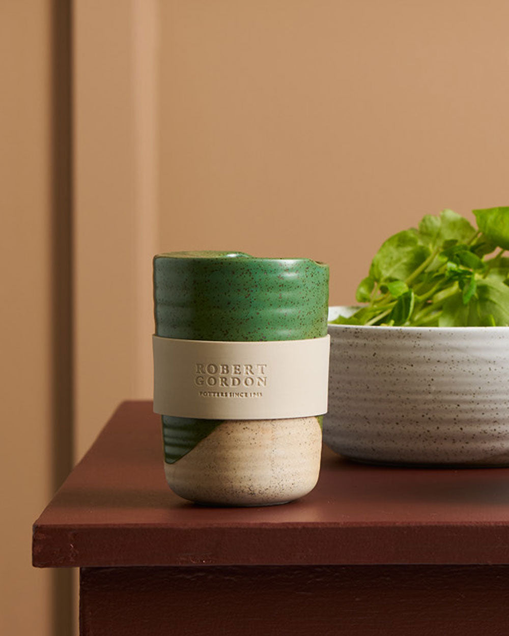 Green travel mug on a burgundy sideboard beside a speckled bowl of herbs
