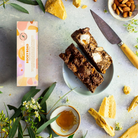 Pebbly Path Honeycomb Highway rocky road 100 g box beside a cut slab on a plate, with honeycomb shards, almonds and honey