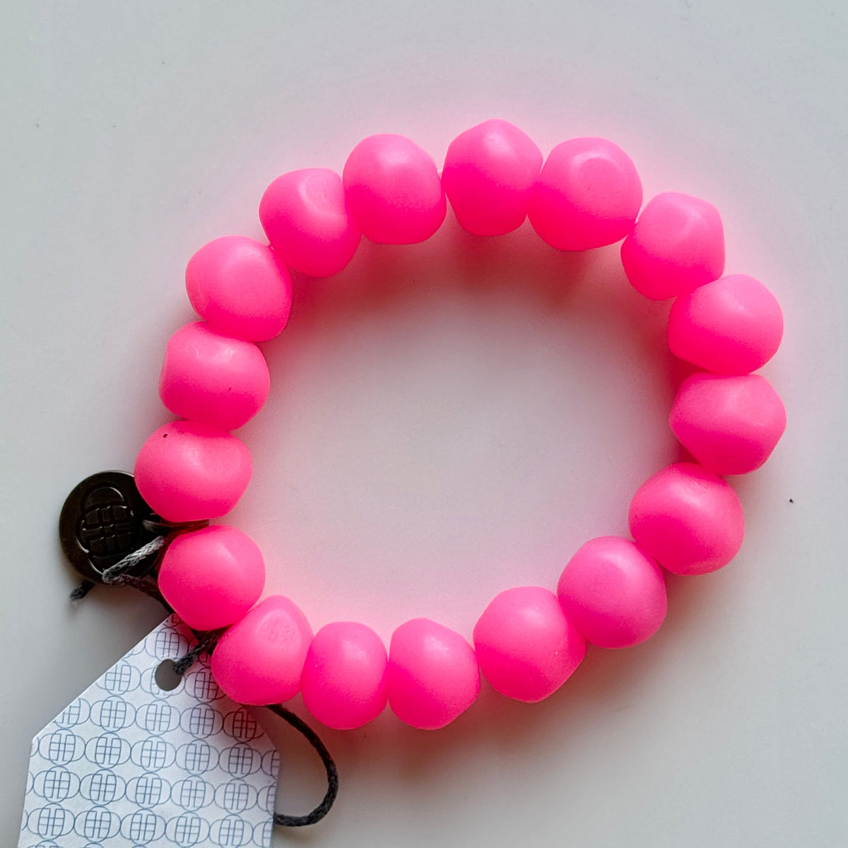 Rock bracelet in Bright Pink: a stretch bracelet of chunky neon pink resin beads with its swing tag and metal charm, laid on a pale surface.