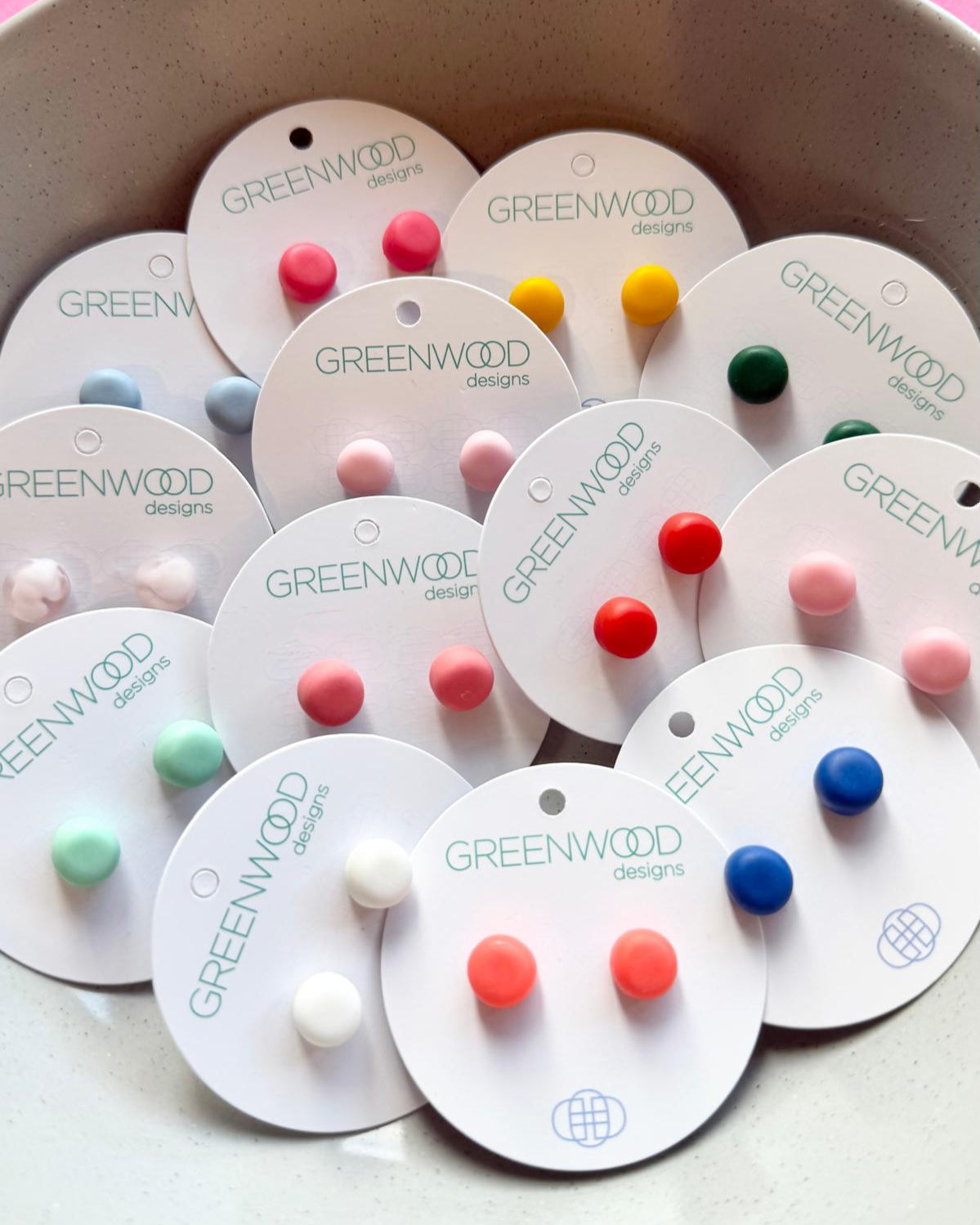 Round resin stud earrings in mixed colours on their round display cards, heaped in a pale stone bowl.