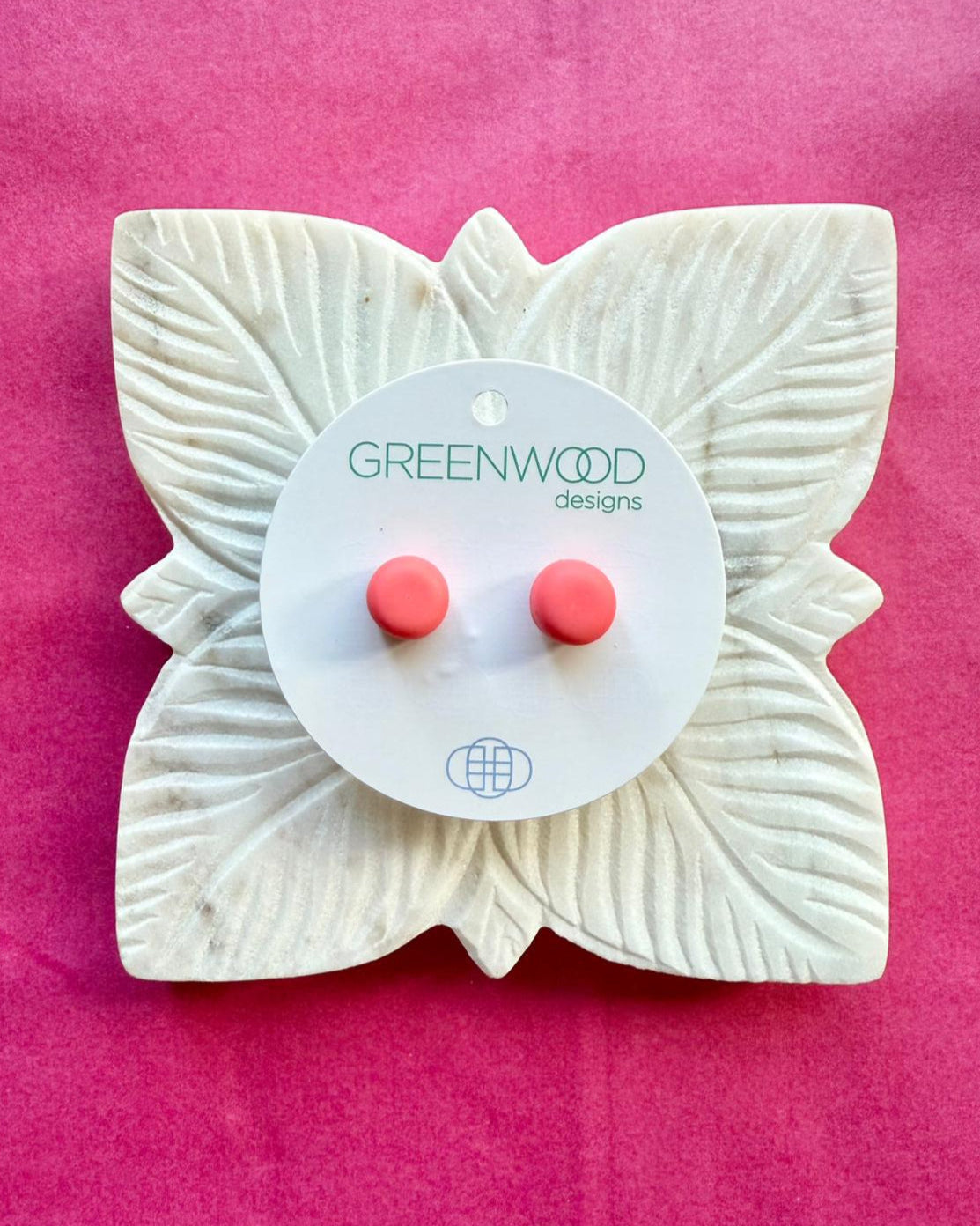 A pair of salmon-coral round resin stud earrings on a round card, on a carved white leaf dish against pink paper.