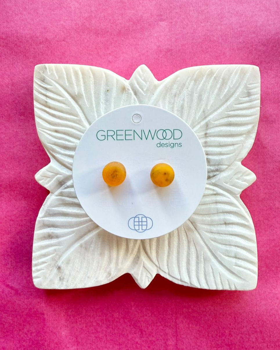 Round earrings in Tortoiseshell: a pair of translucent amber resin studs with darker flecks on a round card, on a carved white leaf dish against pink paper.