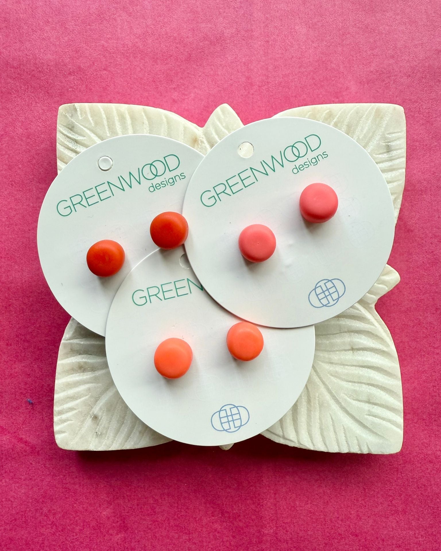 Three pairs of round resin studs in red, coral and orange on overlapping cards, on a carved white leaf dish.