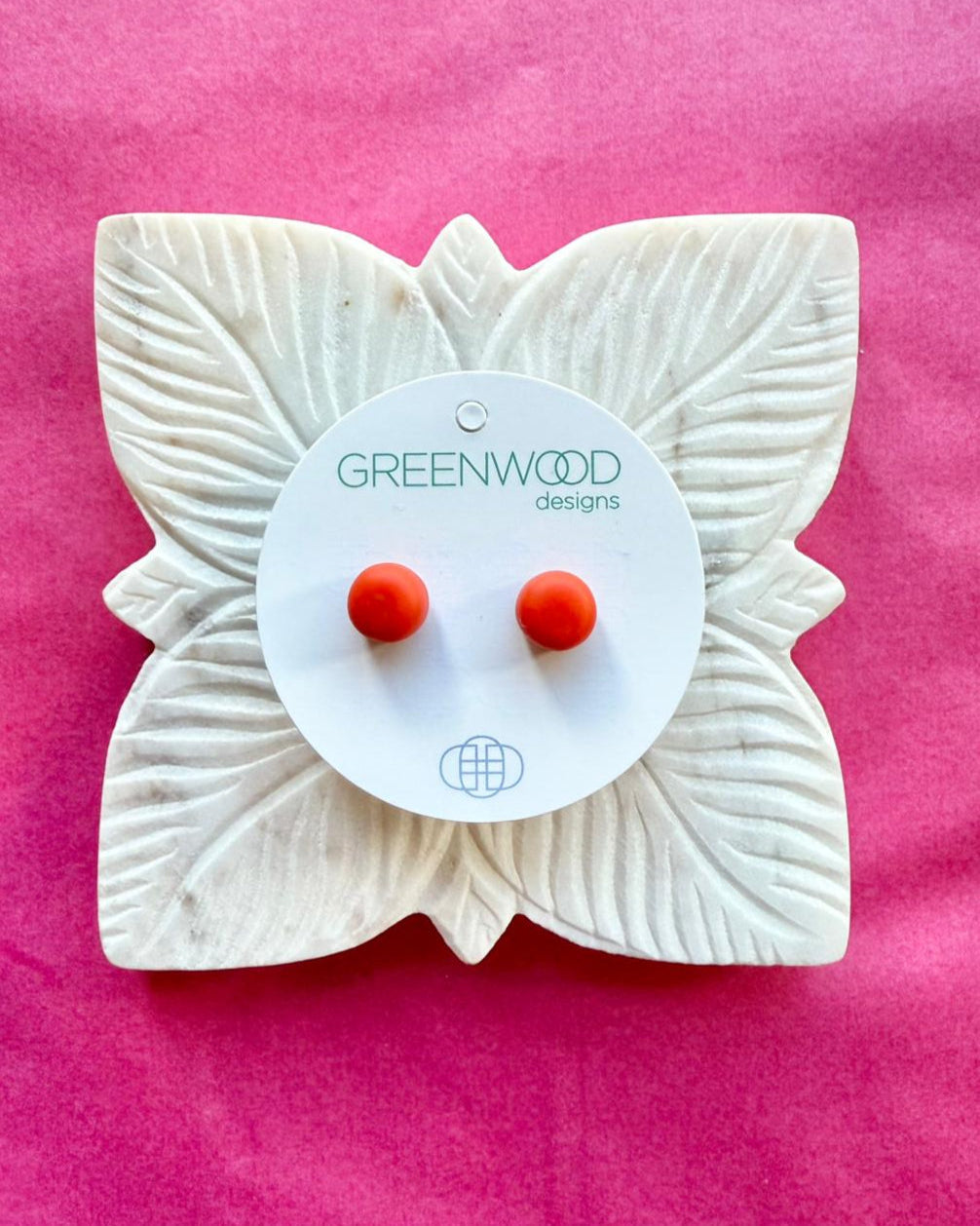 Round earrings in Red: a pair of warm tomato-red resin studs on a round card, on a carved white leaf dish against pink paper.