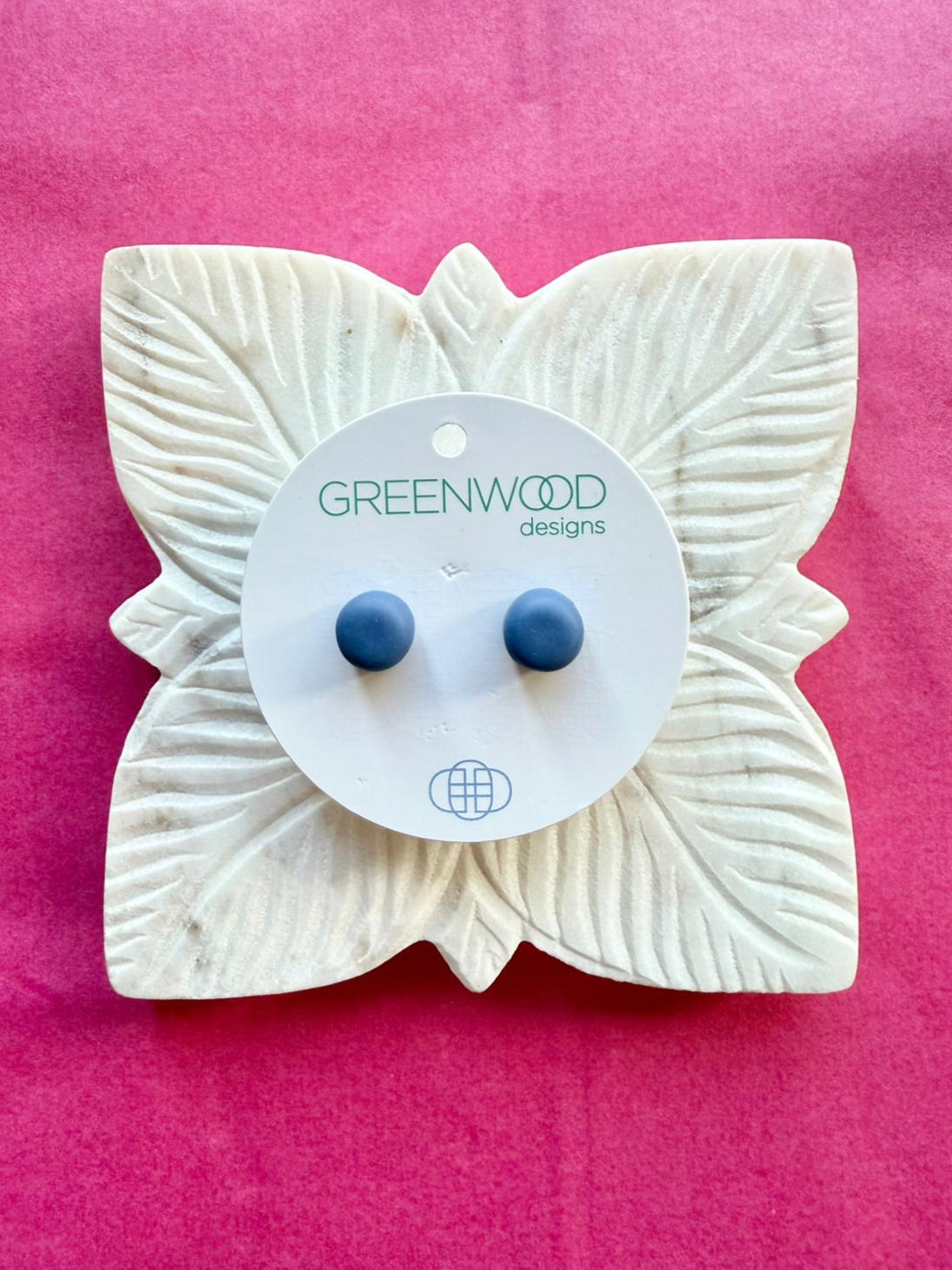 A pair of mid denim-blue round resin stud earrings on a round card, on a carved white leaf dish against pink paper.