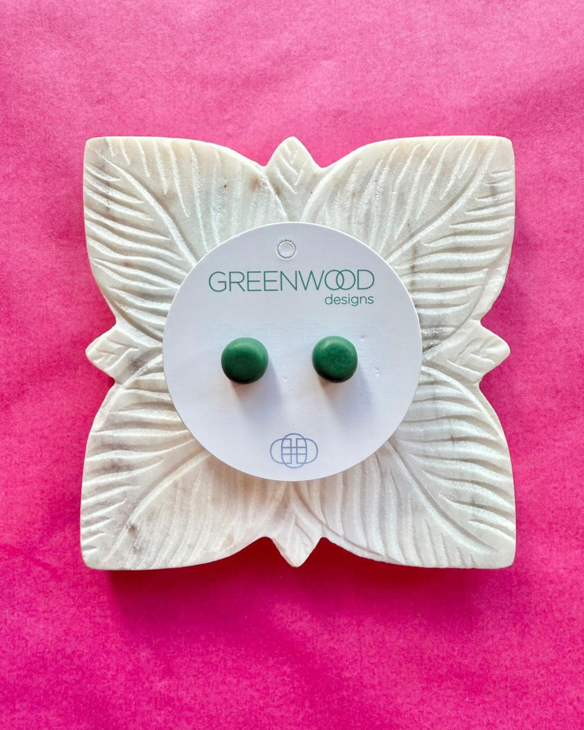 Round earrings in Olive Green: a pair of deep forest-green resin studs on a card, set on a carved white leaf dish against a pink background.