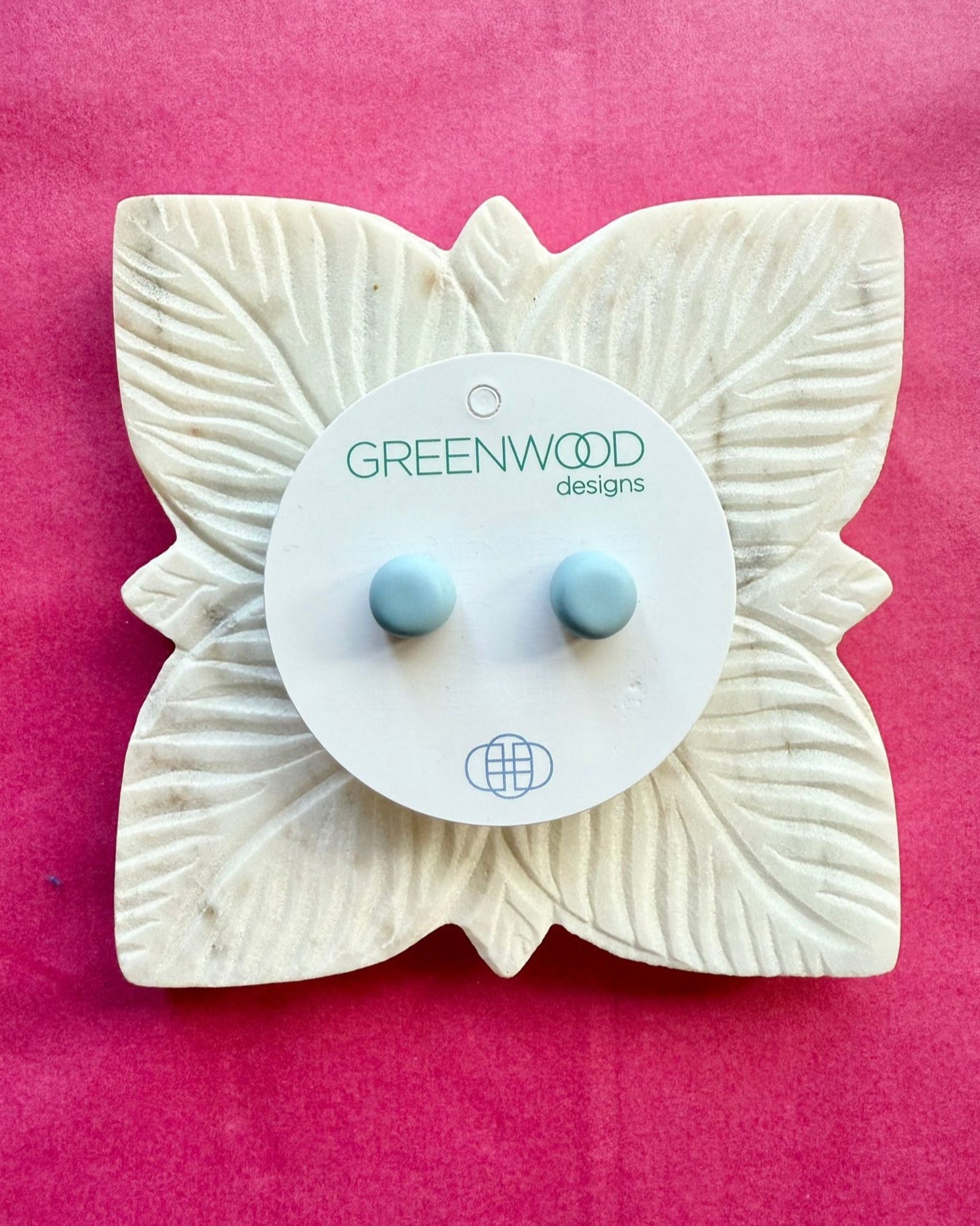 Round earrings in Baby Blue: a pair of pale sky-blue resin studs on a round card, on a carved white leaf dish against pink paper.