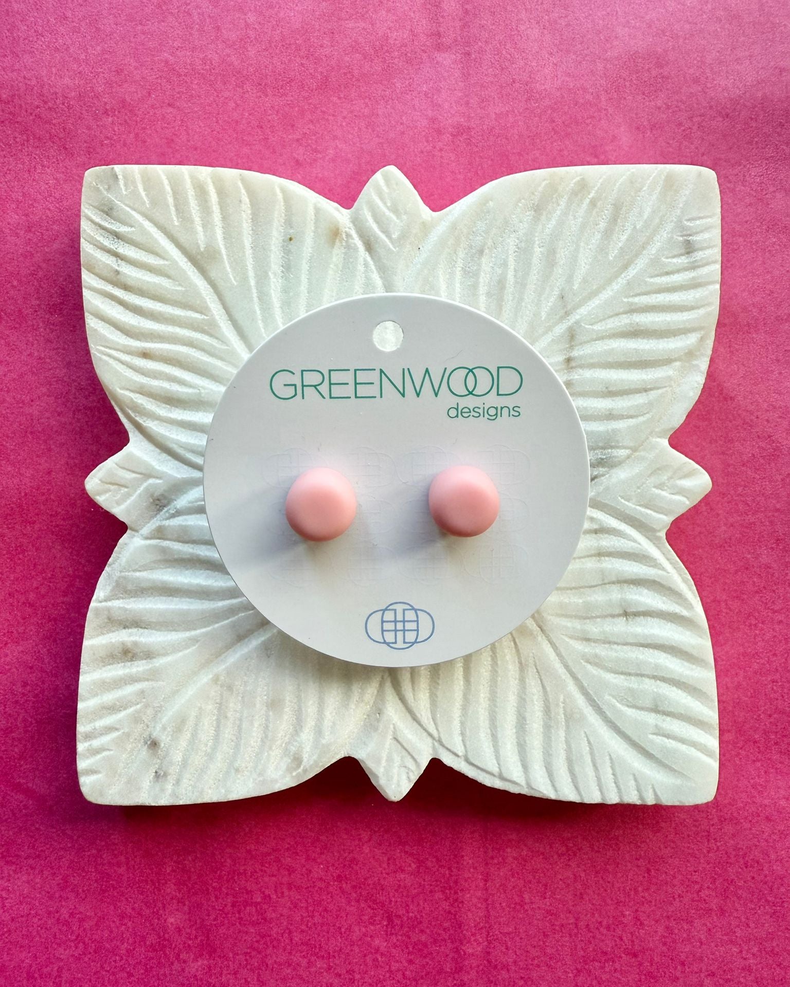 A pair of soft light pink round resin stud earrings on a round card, on a carved white leaf dish against pink paper.