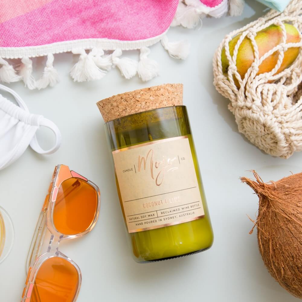 Coconut & Lime candle on a pink tasselled towel with orange sunglasses, a string bag of citrus and a coconut
