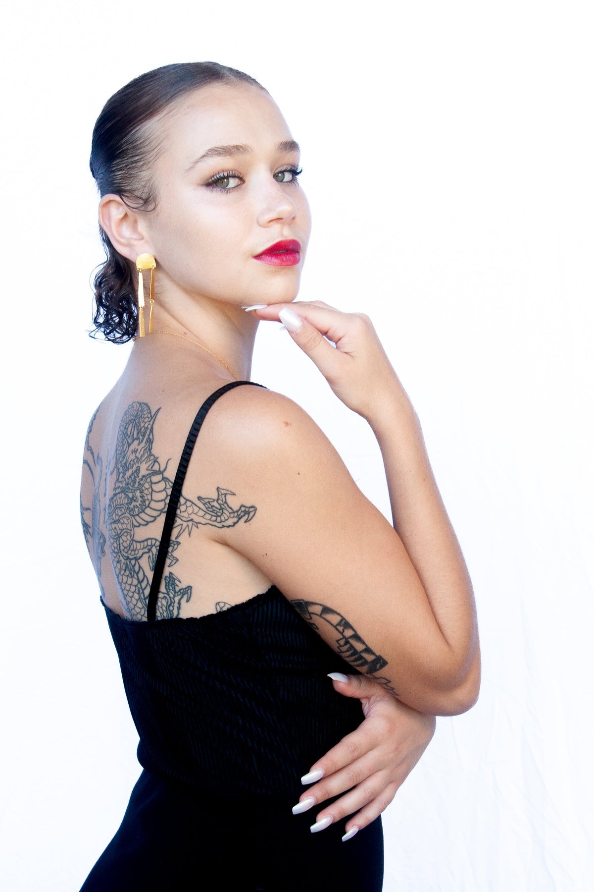Model in a black slip dress, hand at her chin, wearing the gold arch bar-drop earrings