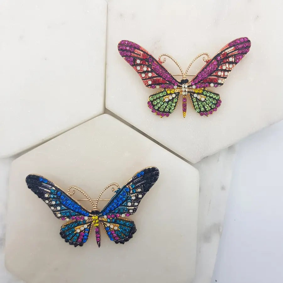 Pink-and-green crystal butterfly brooch beside a blue-and-multicolour one, on marble