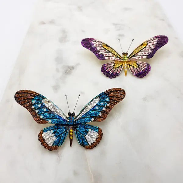 Two crystal butterfly brooches, a purple and yellow one and a blue and brown one, on marble