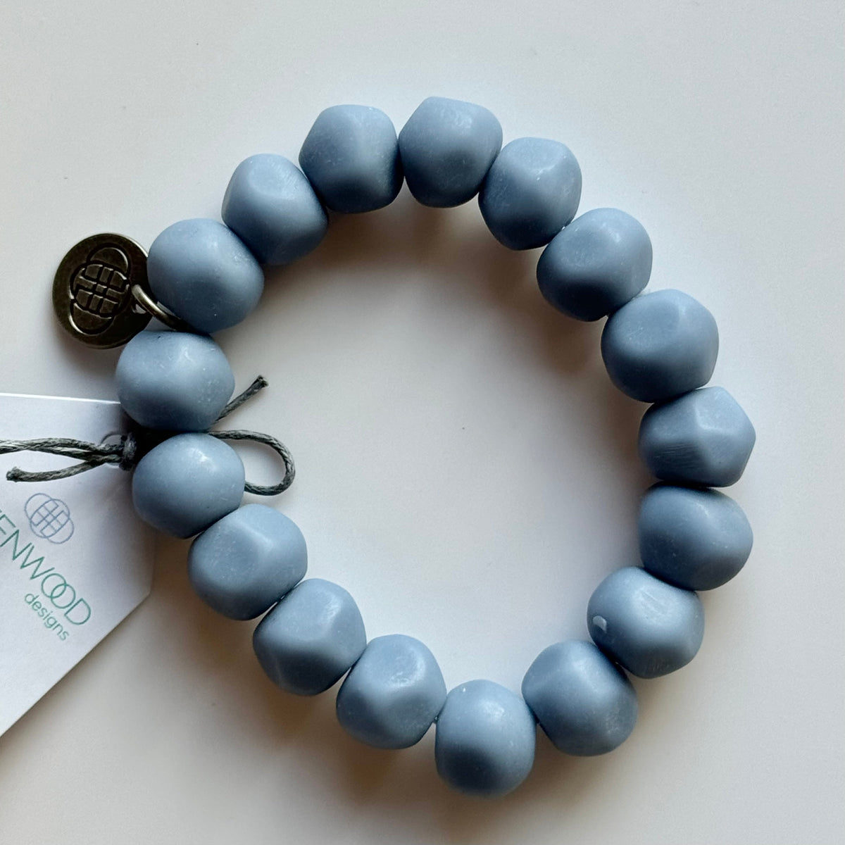 Rock bracelet in Jacaranda: a stretch bracelet of chunky dusty periwinkle-blue resin beads with its swing tag and metal charm, laid on a pale surface.