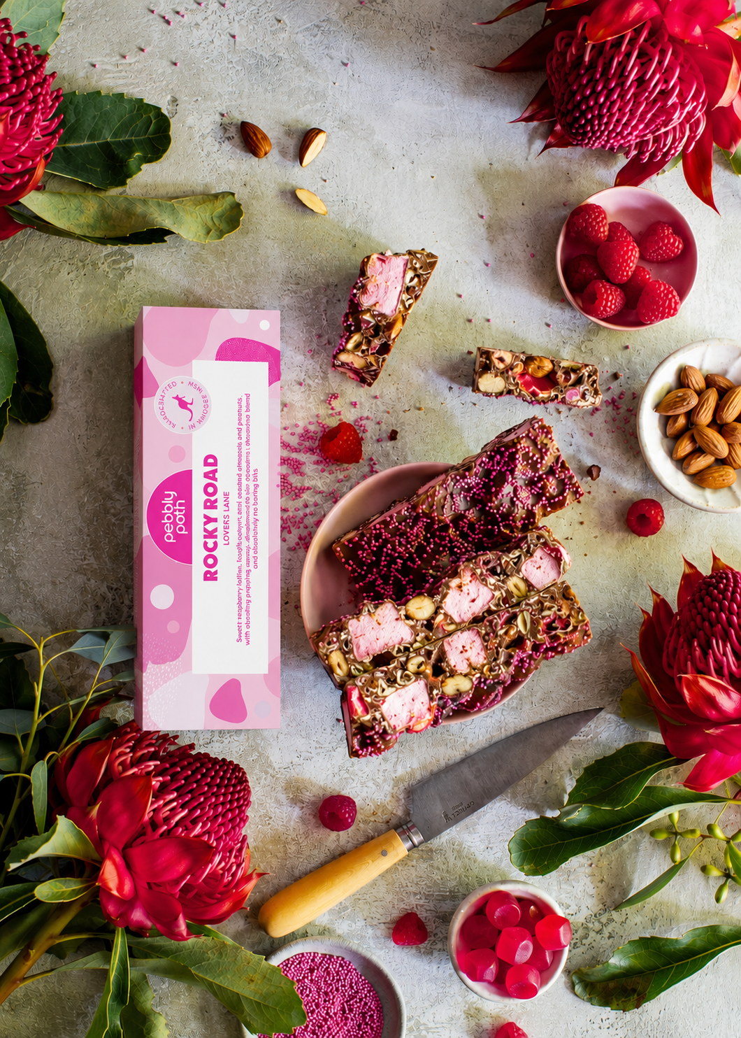 Pebbly Path Lovers Lane rocky road 100 g box with cut slices on a plate, raspberries, almonds and red waratah flowers