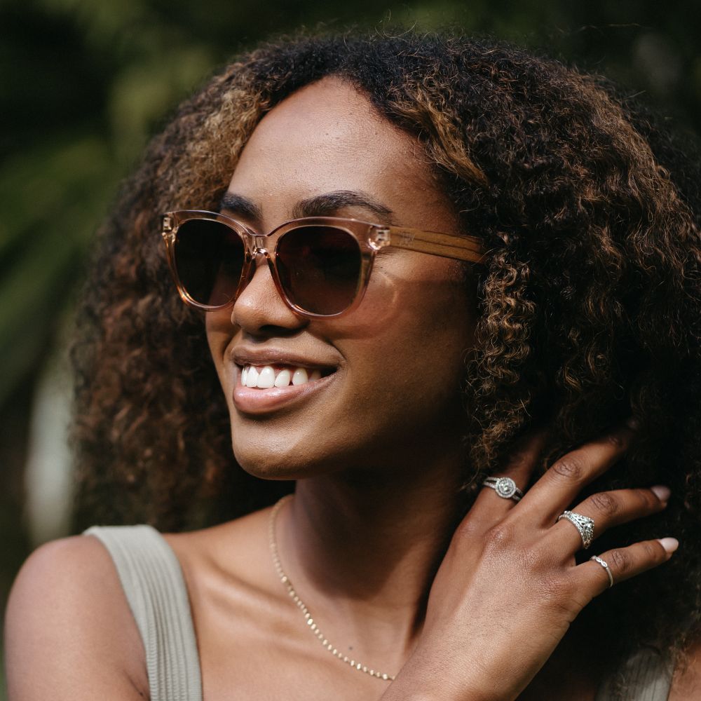 A woman with curly hair smiling outdoors, wearing the Lila Grace sunglasses