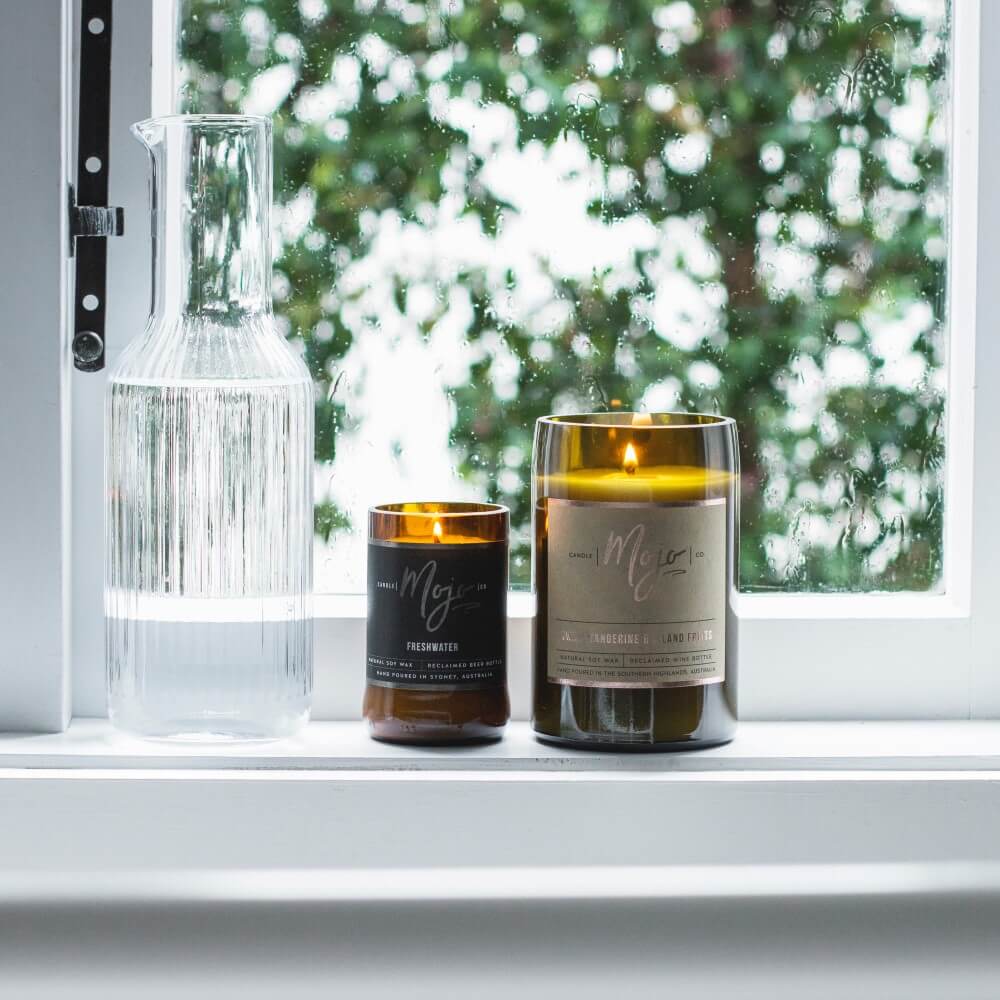 Lit Mojo bottle candles on a sunny windowsill beside a carafe of water