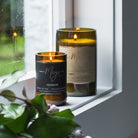 Two lit Mojo bottle candles on a windowsill by a rain-speckled window