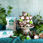 Chunks of Mint Motorway rocky road on a cake stand with marshmallow and green mint jelly, boxes and fresh mint alongside