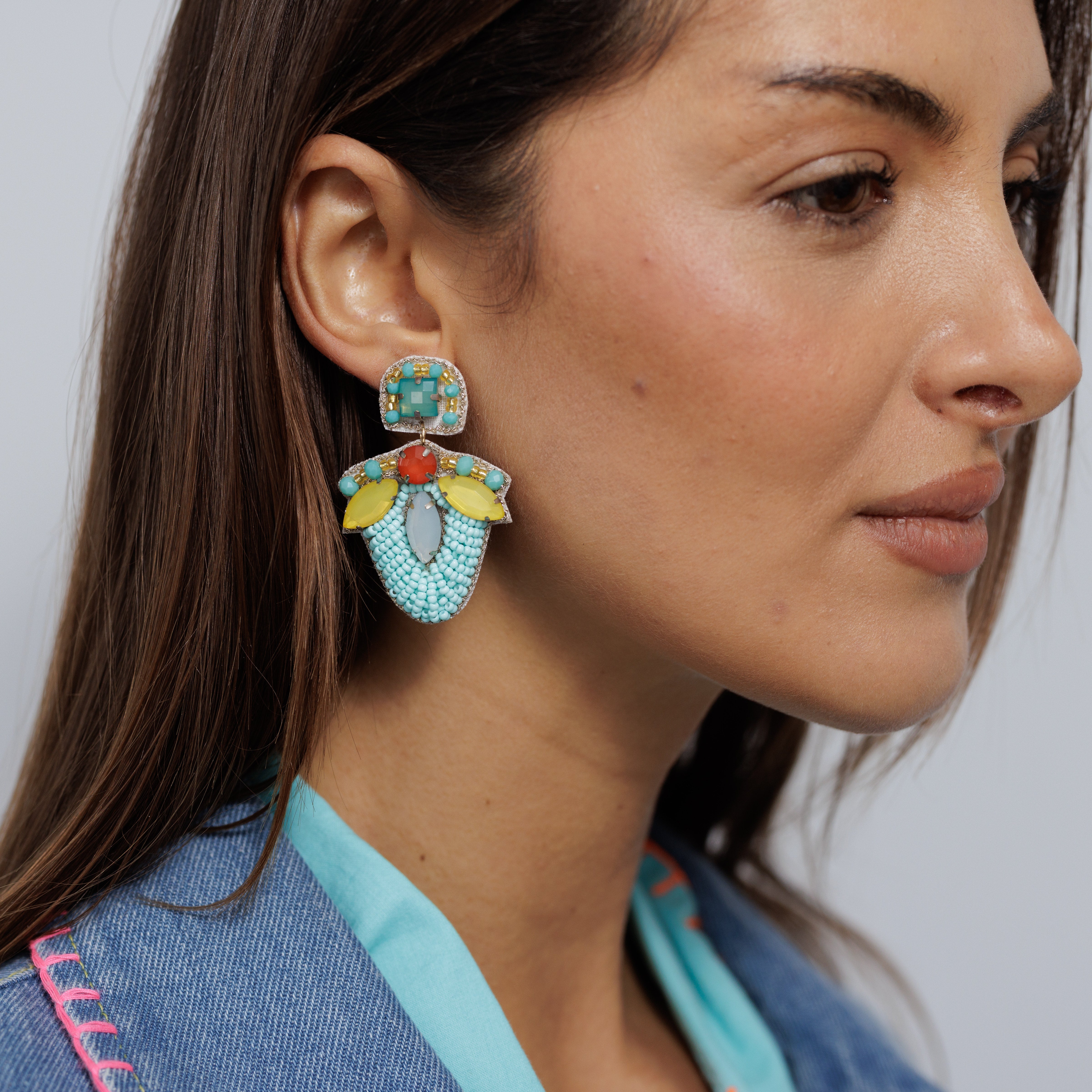 A model wearing a gold-tone drop earring with a pale blue gem stud above a mint and yellow enamel petal.