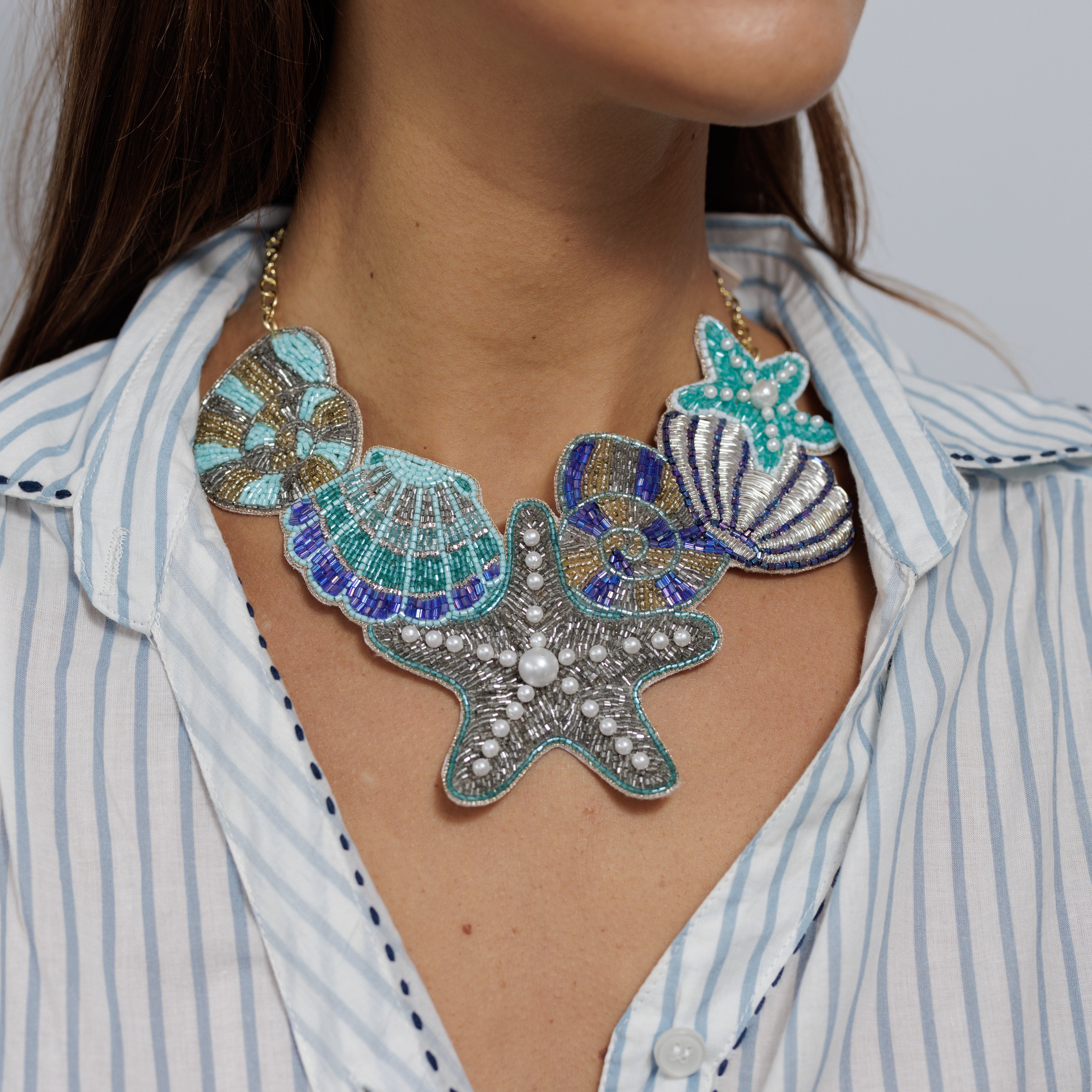 Beaded statement necklace of shells and a starfish in blue, teal and silver, worn over a blue and white striped shirt.