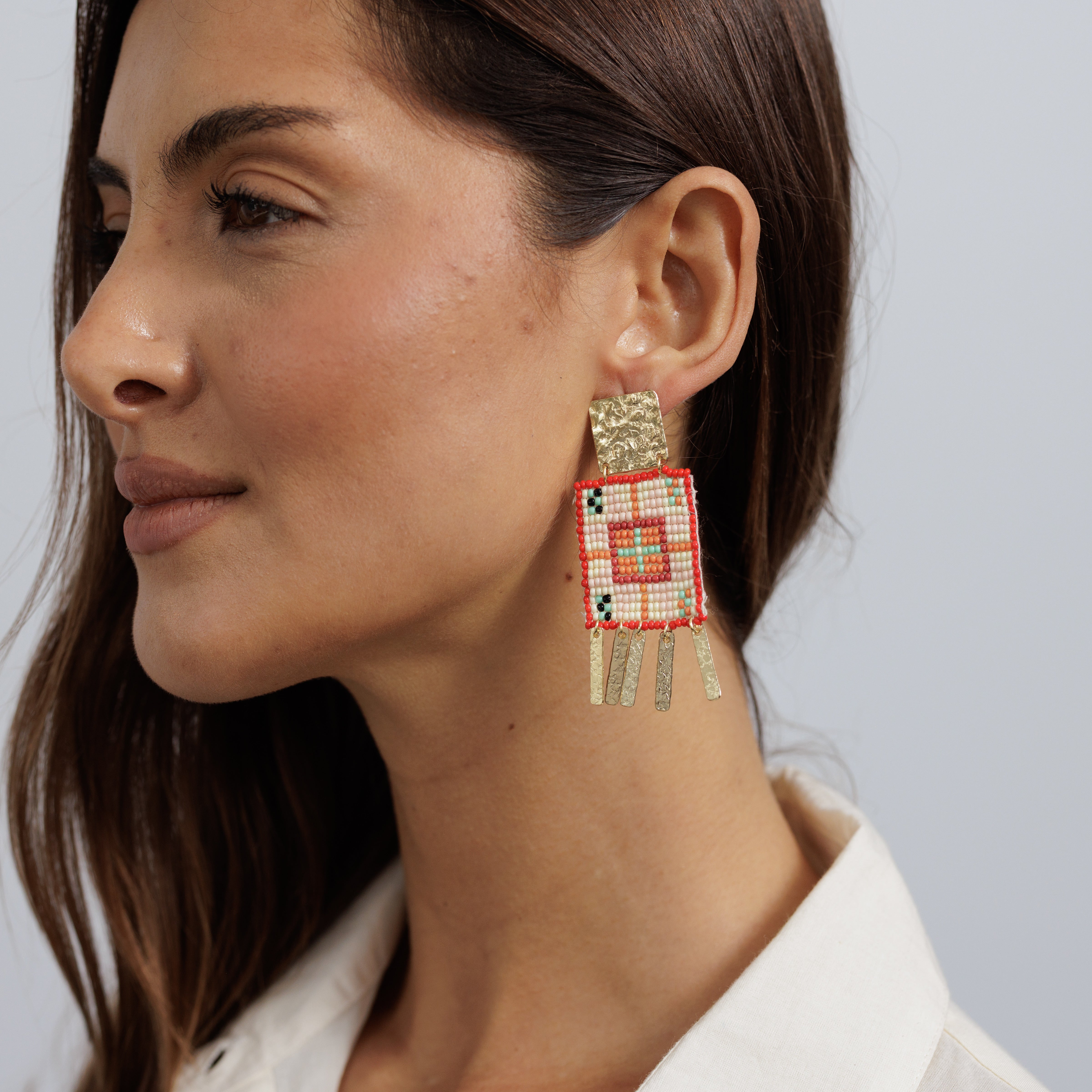 A model wearing a beaded earring with a gold tab above a cream and red woven panel with beaded fringe.