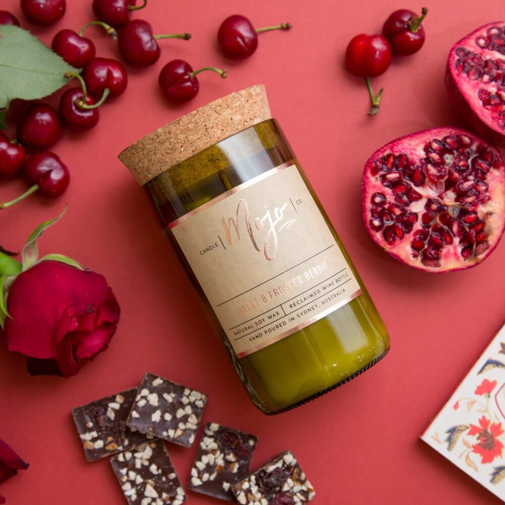 Violet & Frosted Berries candle on a red backdrop with cherries, pomegranate, chocolate and a rose