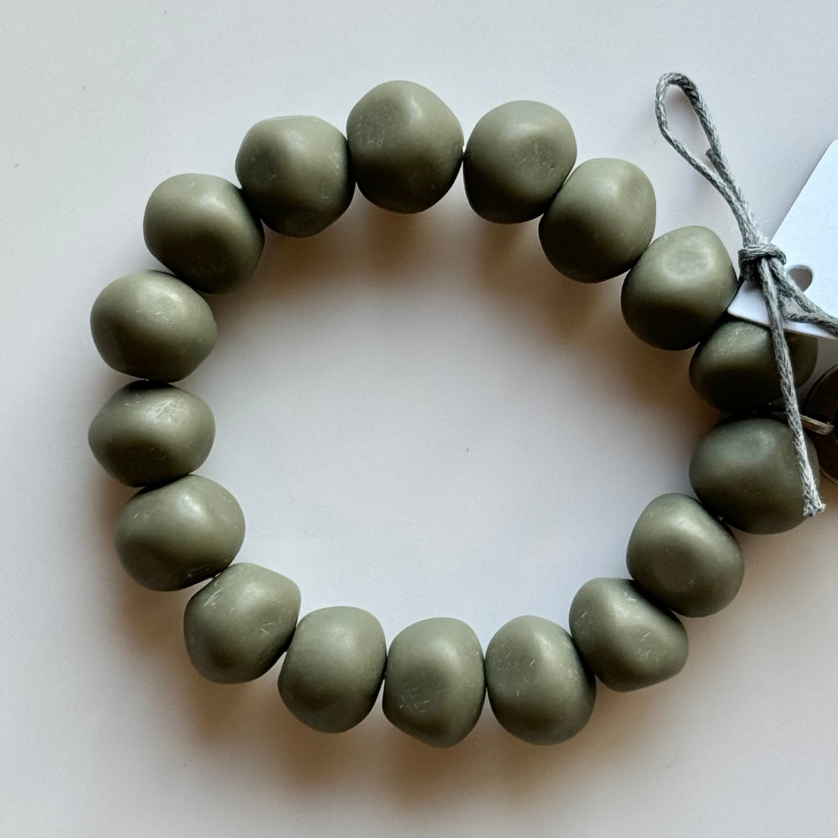 Rock bracelet in Moss: a stretch bracelet of chunky grey-green resin beads with its swing tag and metal charm, laid on a pale surface.