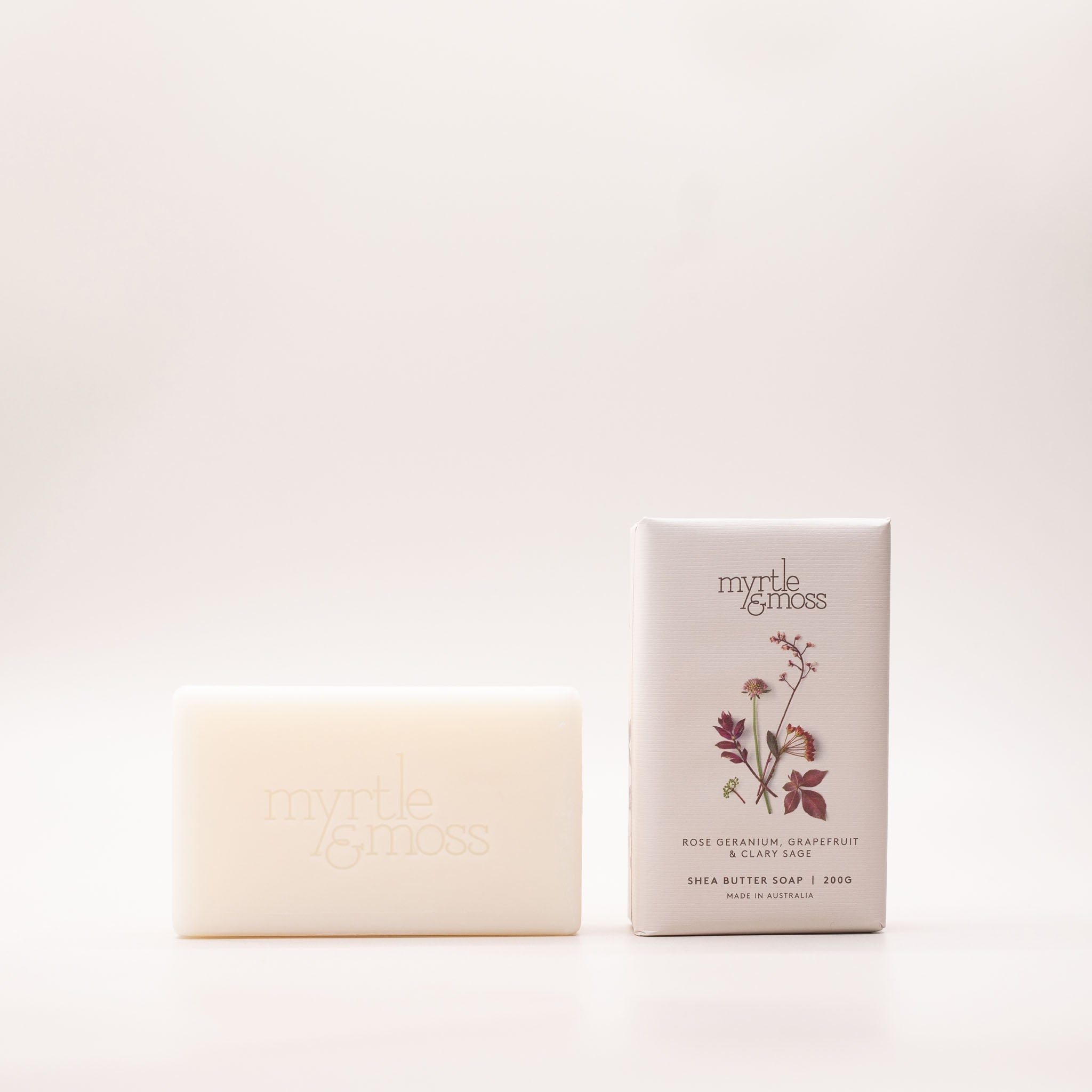 Cream shea butter soap bar beside its pale grey box, printed with deep red and pink pressed blooms and labelled Rose Geranium, Grapefruit & Clary Sage.
