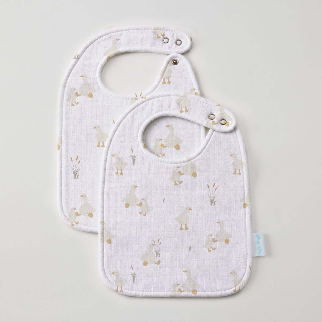 Two white muslin bibs overlapping, printed with small cream ducks and reeds, each with a rounded neck and a press stud at the strap