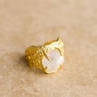 Nohea gold ring with a crushed-metal band holding a raw clear gemstone, on sandstone